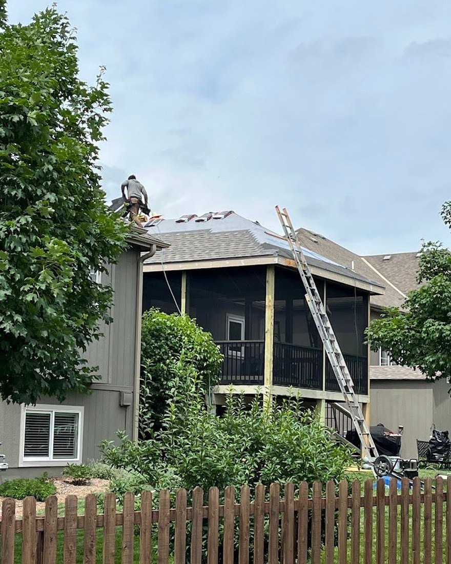 A VCHS crew member laying shingles on a porch roof, with a ladder against the eave.