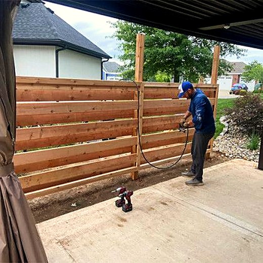Crew member spray-staining a horizontal slat fence.