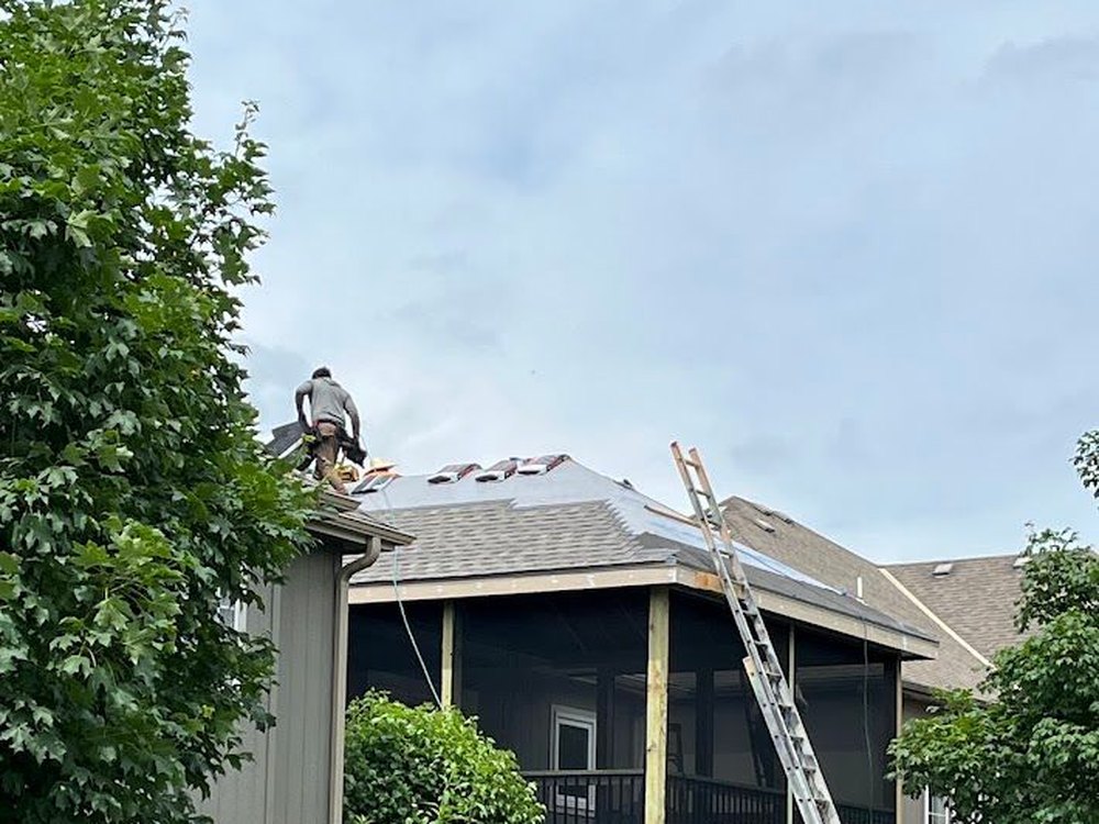 A VCHS crew member laying shingles on a porch roof, ladder against the eave.